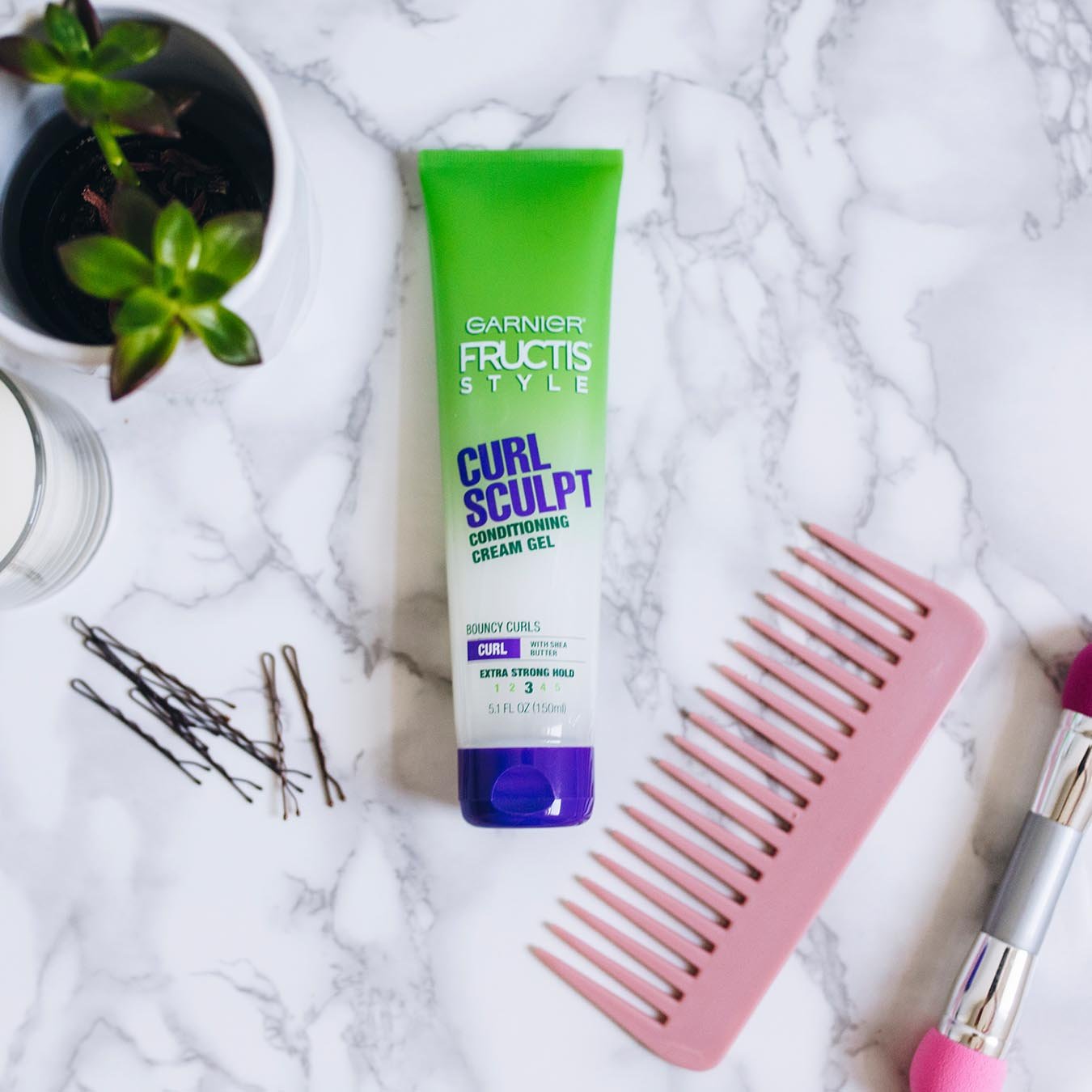 Garnier Fructis Style Curl Sculpt Conditioning Cream Gel on white marble next to bobby pins, a pink comb, a dual-tipped smudging brush, a white candle, and a potted plant.